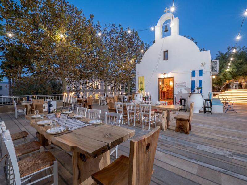 Widok wieczorny na małą białą kapliczkę z drewnianym tarasem i stołami na zewnątrz pod drzewami i lampkami.