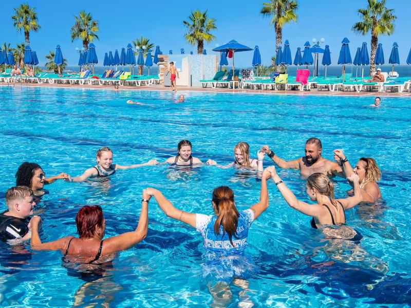 Ludzie tworzą krąg, trzymając się za ręce w basenie, otoczeni palmami i parasolami.