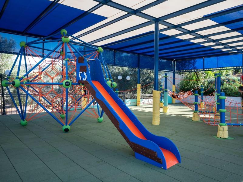 Zadaszony plac zabaw z niebiesko-czerwoną zjeżdżalnią i konstrukcją do wspinaczki w otoczeniu hotelu