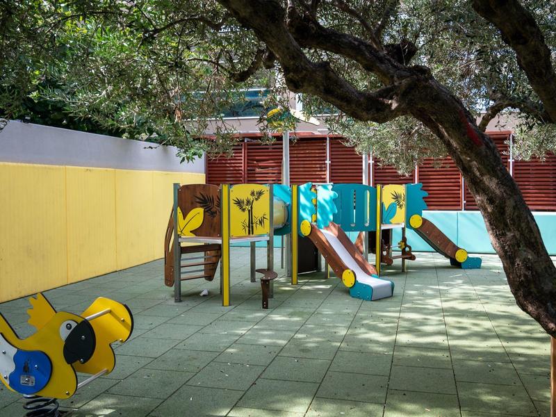 Plac zabaw z zjeżdżalniami i huśtawkami otoczony drzewami i budynkami.