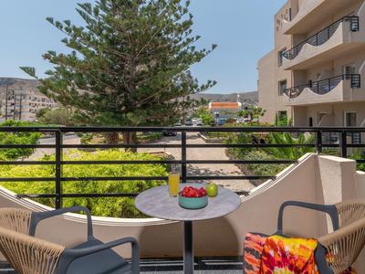 Balcón con dos sillas, una mesa con un plato de frutas y vista a un árbol y edificio del hotel.