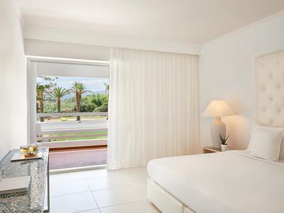 Jasny pokój hotelowy z białym łóżkiem, zasłonami i widokiem na taras z palmami.