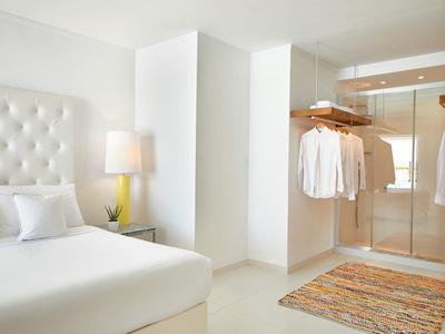 Jasna sypialnia z białym łóżkiem, stolikiem nocnym, lampą i otwartą szafą z koszulami.