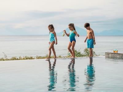 Troje dzieci w strojach kąpielowych balansuje na krawędzi basenu typu infinity z widokiem na morze.