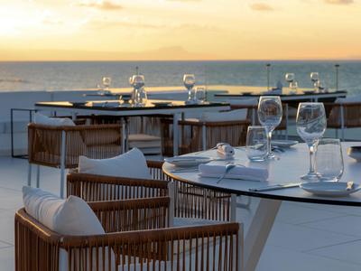 Elegante terrazza esterna con tavoli apparecchiati e sedie in legno sul mare al tramonto.