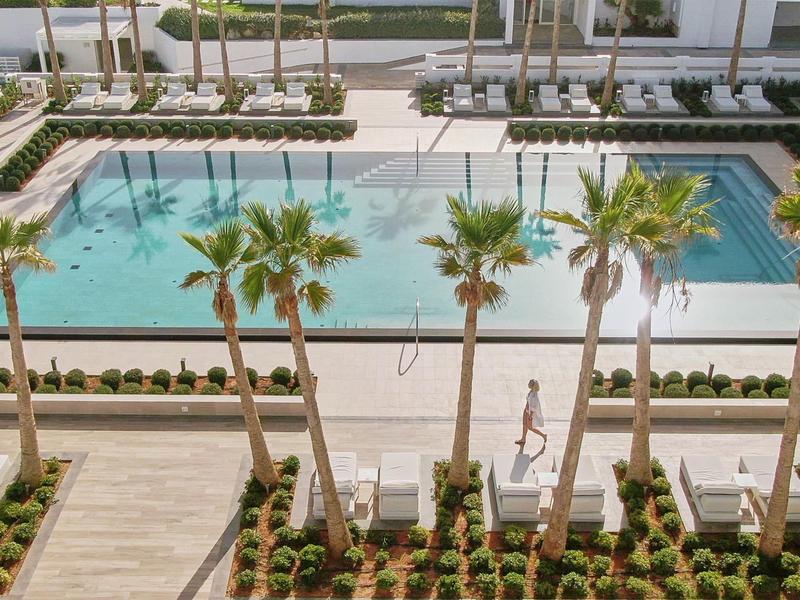 Widok na zadbany basen z palmami i leżakami obok hotelu.
