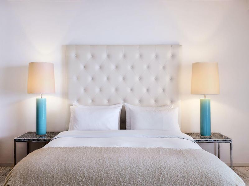 Nowoczesne łóżko podwójne z pikowanym zagłówkiem, białymi poduszkami i dwoma niebieskimi lampami na stolikach nocnych.
