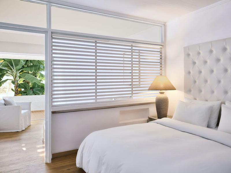 Nowoczesny pokój hotelowy z białym łóżkiem, pikowanym zagłówkiem i widokiem na taras z rośliną.