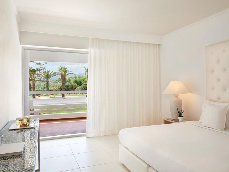 Jasny pokój hotelowy z białym łóżkiem, zasłonami i widokiem na taras z palmami.