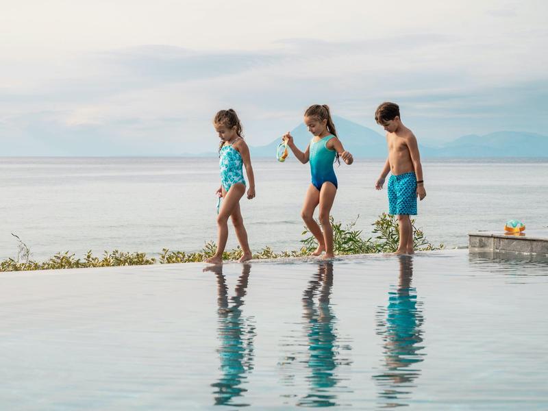 Troje dzieci w strojach kąpielowych balansuje na krawędzi basenu typu infinity z widokiem na morze.