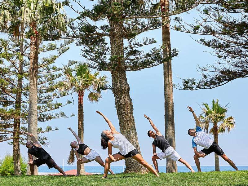 Pięć osób ćwiczy jogę na świeżym powietrzu między palmami a sosnami pod jasnym niebem.