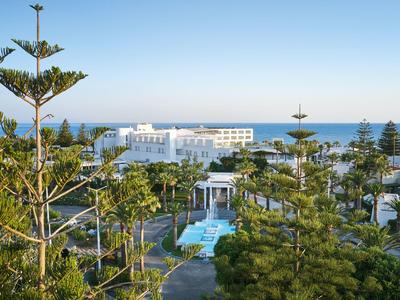 Vista de un hotel con piscina, rodeado de árboles y vista al mar al fondo.