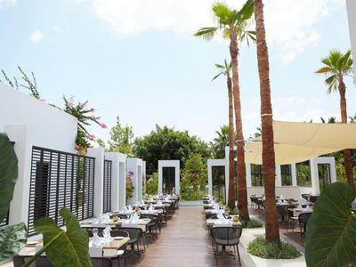 Área exterior de un restaurante moderno con mesas, sillas y palmeras bajo un cielo azul.