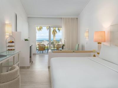 Habitación de hotel luminosa con cama doble, zona de estar y balcón con vistas al mar.