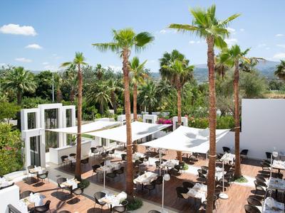 Restauracja na świeżym powietrzu z białymi stołami, krzesłami i daszkami w otoczeniu palm i gór.