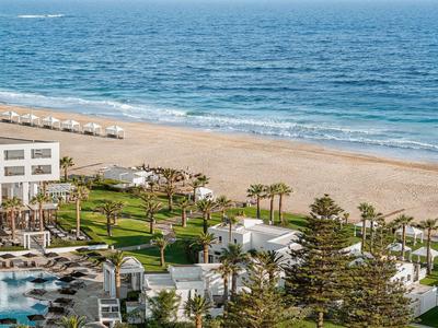 Widok na basen hotelowy i zadbane ogrody na plaży z niebieskim morzem w tle.