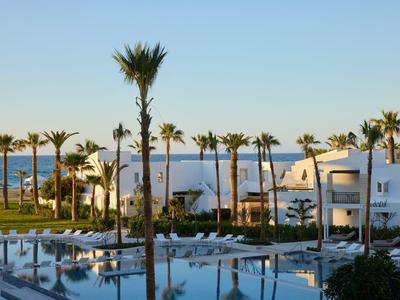 Hotel bianco con piscina e palme vicino a una spiaggia soleggiata.