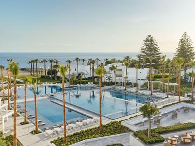 Grande piscina esterna con palme e vista mare, circondata da sdraio e edifici bianchi.