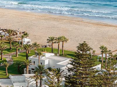 Complesso alberghiero moderno con edifici bianchi, palme e vista su una vasta spiaggia di sabbia e il mare.