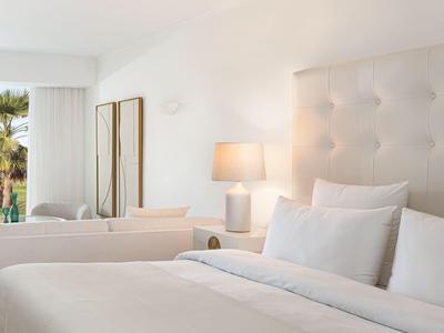 Camera d'hotel luminosa con letto bianco, testiera capitonné e lampada da tavolo accanto al divano.