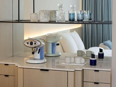 Bancone moderno in camera d'hotel con macchina per caffè, bicchieri e tazze davanti a letto con cuscini bianchi.