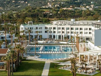 Hotel moderno con grande piscina, lettini e circondato da palme in un paesaggio verde.