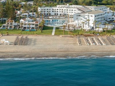 Grande hotel sulla spiaggia con piscina, lettini e costa marina blu in primo piano.