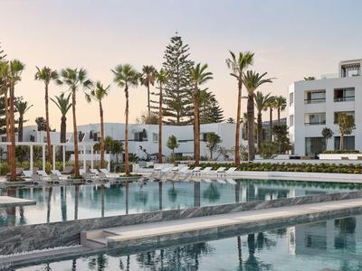 Piscina moderna di un hotel con lettini e palme davanti a un edificio bianco al tramonto.