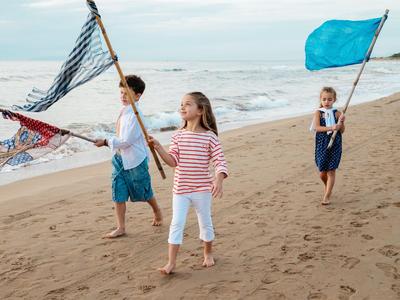 Tre bambini con bandiere camminano lungo la spiaggia con il mare sullo sfondo.