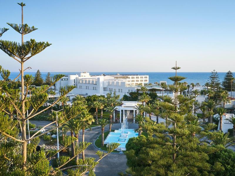 Vista de un hotel con piscina, rodeado de árboles y vista al mar al fondo.