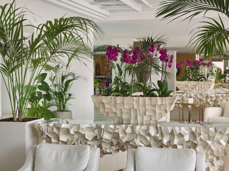 Zona luminosa del hotel con muebles blancos, plantas verdes y orquídeas moradas sobre mesas decorativas.