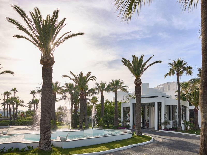 Hotel con edificio blanco rodeado de palmeras y una piscina al atardecer.