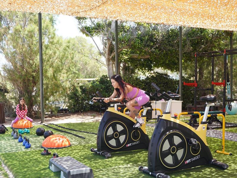 Mujer entrenando en una bicicleta estática al aire libre bajo una sombra, rodeada de plantas.