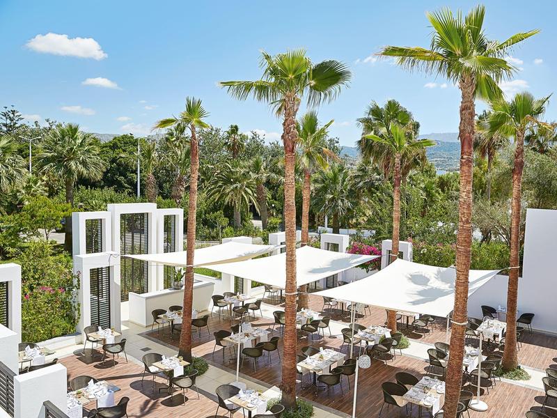 Zona al aire libre de hotel con muebles acolchados blancos y palmeras bajo un cielo azul