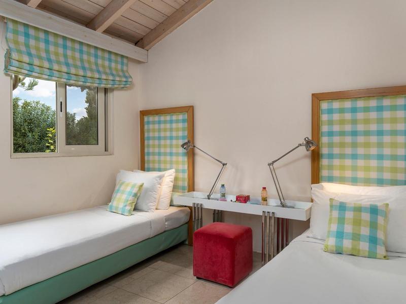 Dos camas individuales con cabeceras a cuadros, ropa blanca y taburetes rojos en una habitación luminosa.