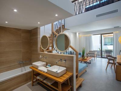 Chambre d'hôtel moderne avec baignoire, deux lavabos, coin salon et balcon avec vue sur la mer.