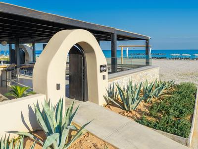 Modern hotel entrance with white arch, plants, and direct beach view under clear sky.