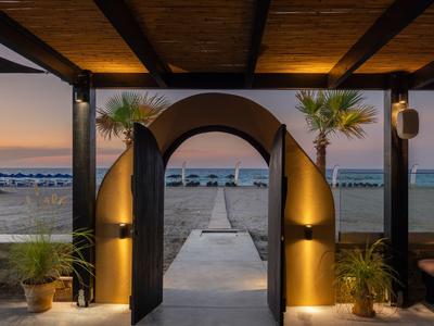 View through an arch to a pier extending to the sea at sunset with illuminated plants in a hotel area.