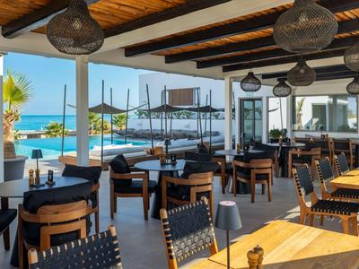 Covered outdoor hotel area with wooden tables and chairs, overlooking pool and sea.