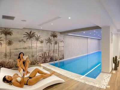 Two women relax on loungers beside an indoor pool in a stylish wellness area.
