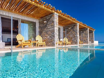 Hébergement de luxe avec piscine à débordement et chaises longues devant une terrasse en pierre sous un ciel bleu.
