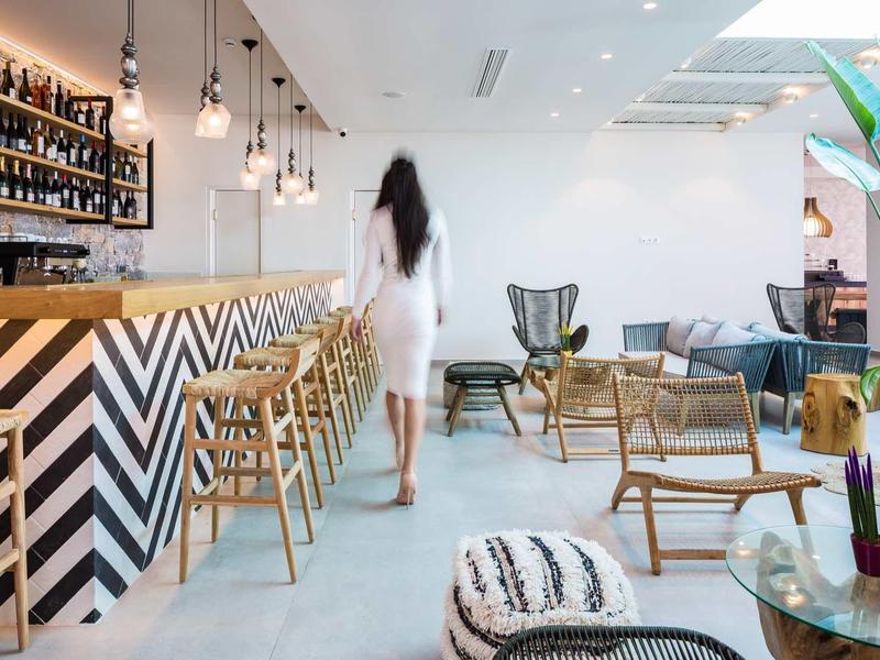 Café d'hôtel moderne avec bar en bois, tabourets, coin salon confortable avec plantes et tapis.