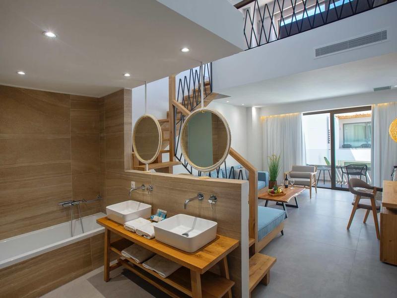 Chambre d'hôtel moderne avec baignoire, deux lavabos, coin salon et balcon avec vue sur la mer.