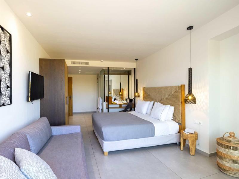 Chambre d'hôtel lumineuse avec lit double, canapé gris, TV murale et décoration moderne.