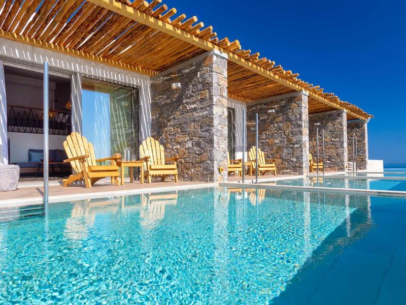 Hébergement de luxe avec piscine à débordement et chaises longues devant une terrasse en pierre sous un ciel bleu.