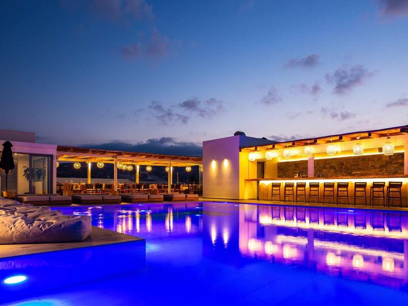 Piscine et zone de bar éclairées dans un hôtel au coucher du soleil.