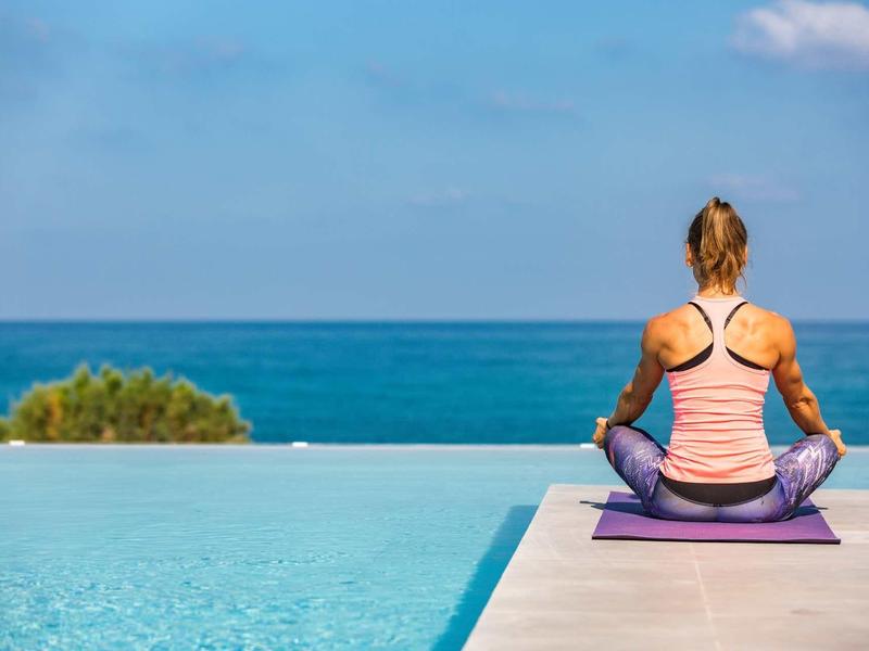 Femme méditant sur tapis de yoga au bord d'une piscine à débordement avec vue sur la mer.