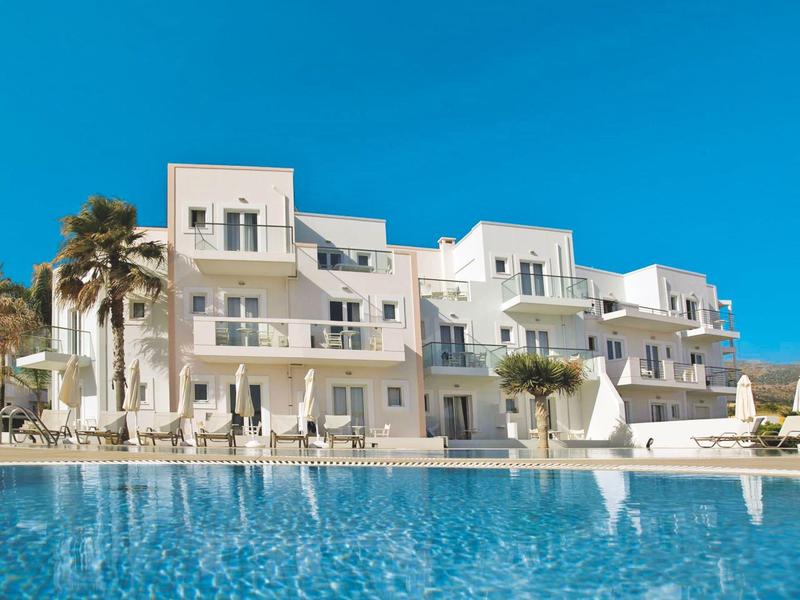 Moderní bílá hotelová budova s velkými okny vedle modrého bazénu pod jasnou oblohou.