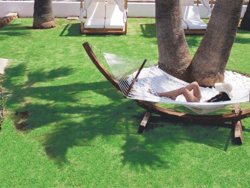 Osoba odpočívající na houpací síti na zahradě s palmami a trávou.