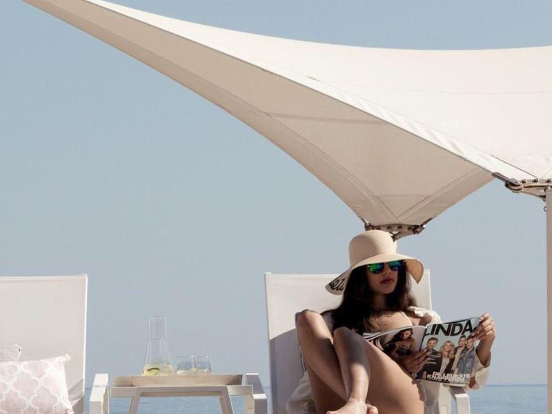 Žena odpočívající na židli pod slunečníkem na pláži vedle malého stolku s nápoji.